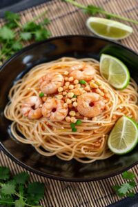 Pad thai near me servido em prato escuro com camarões, amendoim crocante, broto de feijão e limão, textura brilhante do molho tailandês