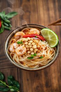 Pad thai recipe tradicional com macarrão de arroz, camarões, tofu e amendoim em bowl cerâmico, decorado com limão e pimenta