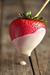 Morango do amor pronto: morango coberto com recheio rosa e mergulhado em chocolate branco brilhante, espetado em palito.