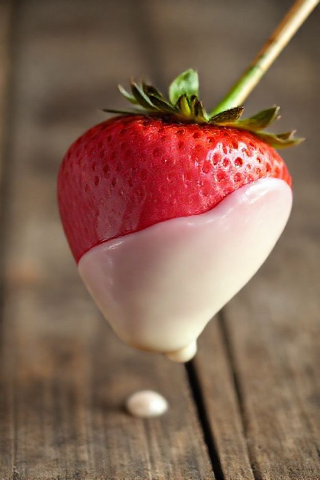 Morango do amor pronto: morango coberto com recheio rosa e mergulhado em chocolate branco brilhante, espetado em palito.